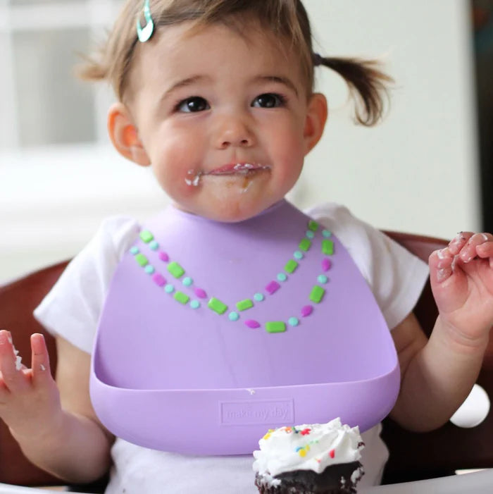 Make My Day Baby Bib Lilac Jewel worn by a toddler, showcasing a lilac silicone bib with colorful bead pattern and frosting on mouth.
