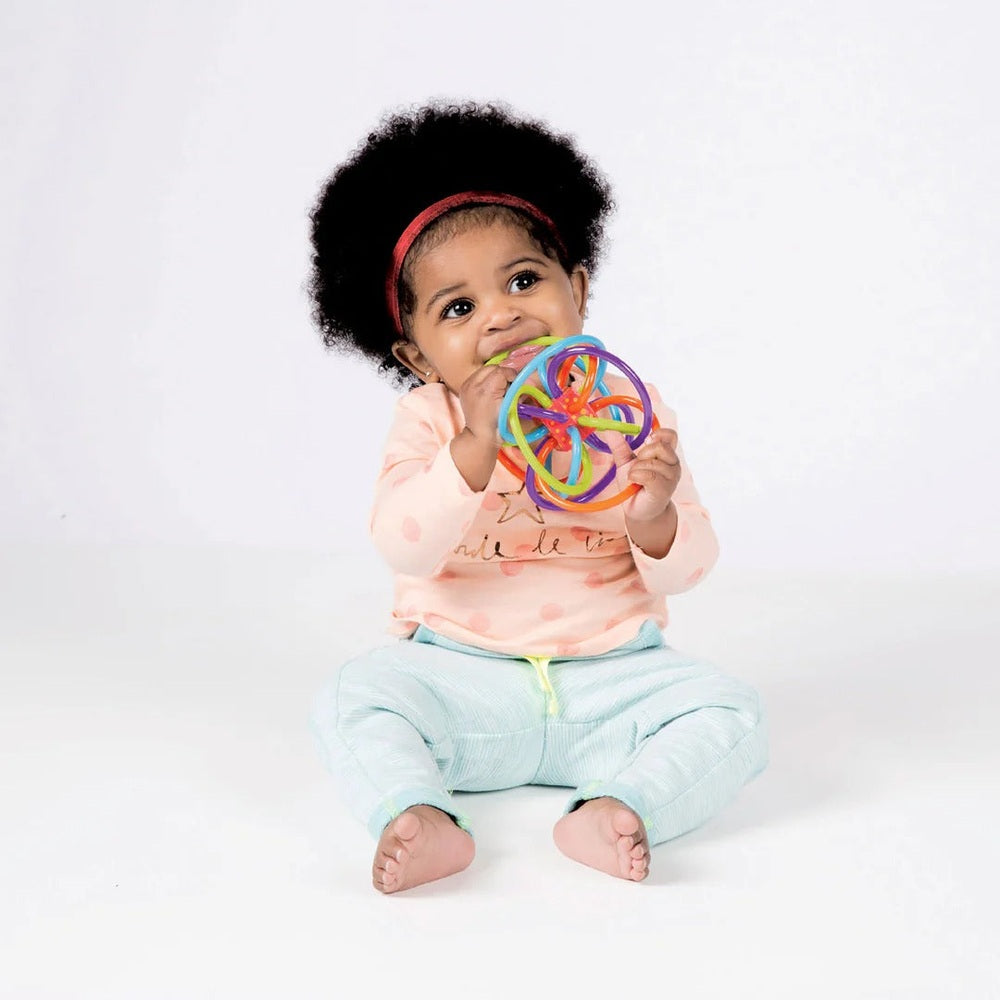 Manhattan Toy Winkel Rattle Sensory Teether Toy in rainbow colors; baby sits and chews the toy.