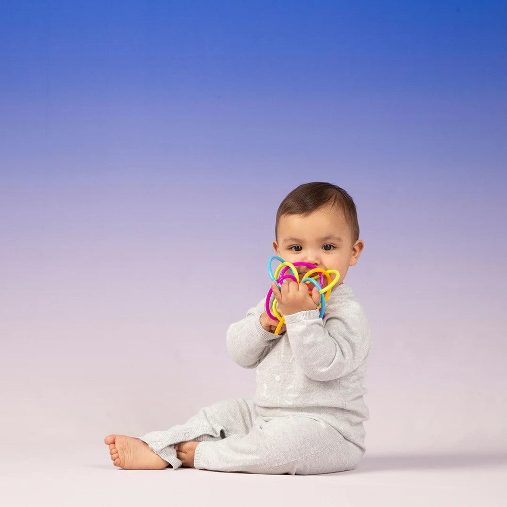 Manhattan Toy Winkel Rattle Sensory Teether Toy; baby sits in gray pajamas, chewing colorful rings.