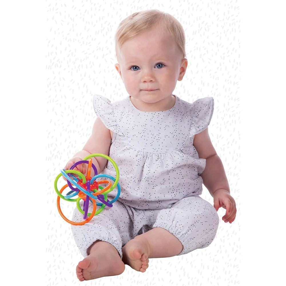 Manhattan Toy Winkel Rattle Sensory Teether Toy held by a baby in a white speckled outfit with colorful rings.