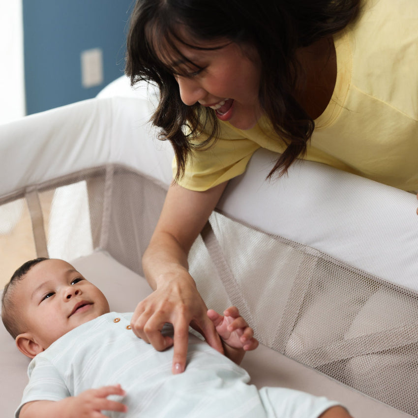 MAXI COSI Swift Playard bassinet; white mesh sides, baby lying down as caregiver reaches in.