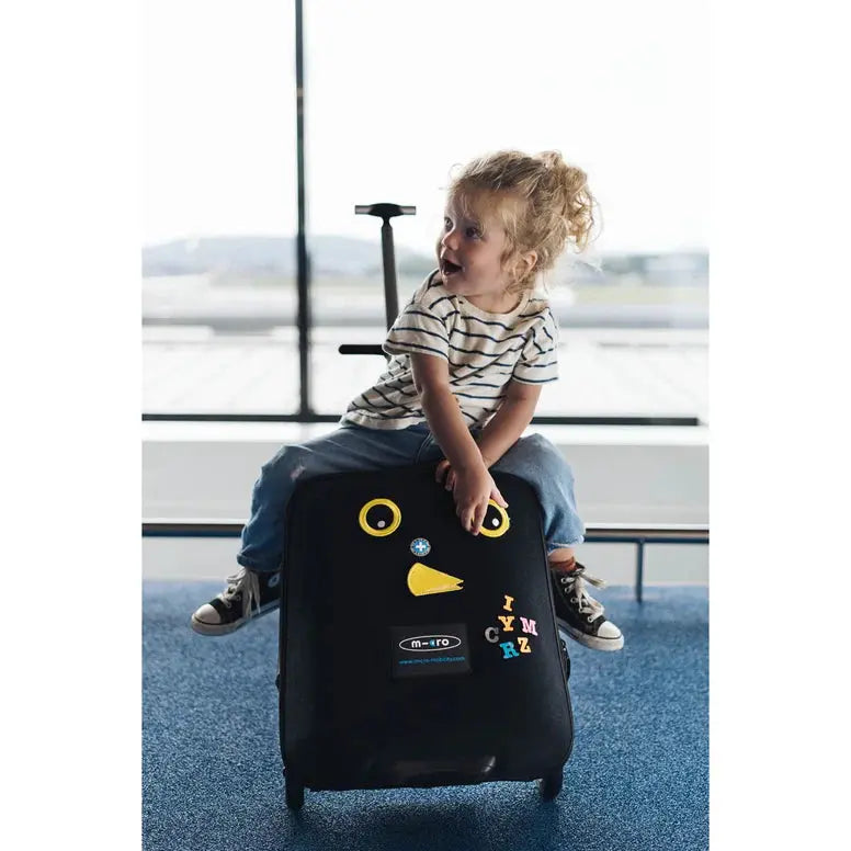 Micro Eazy Luggage suitcase in black with yellow eyes and beak, child sits on it at the airport.