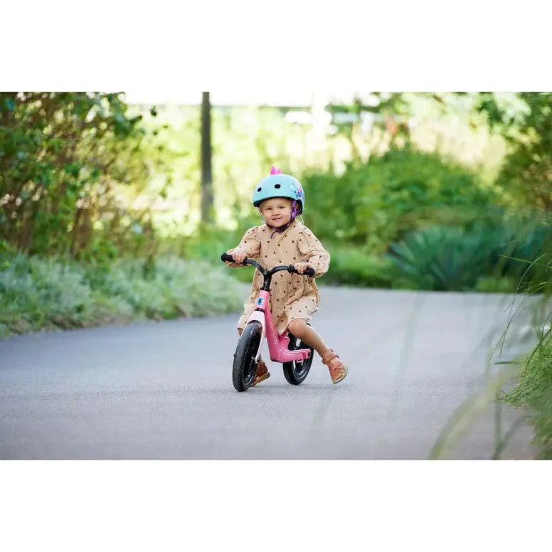 Micro Helmet V2 Dragon toddler rides a pink balance bike on a sunny outdoor path, wearing a blue helmet.