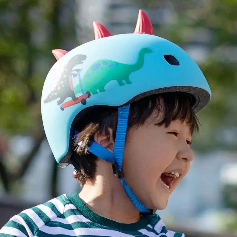 Micro Helmet V2 Scootersaurus blue helmet with dinosaur print; child fastens the blue chin strap while laughing.