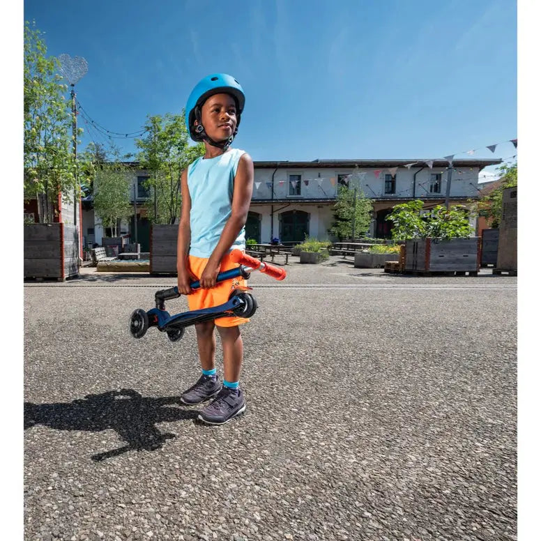 Micro Maxi Foldable Deluxe LED Scooter, blue frame with orange handlebars, carried by a helmeted child.
