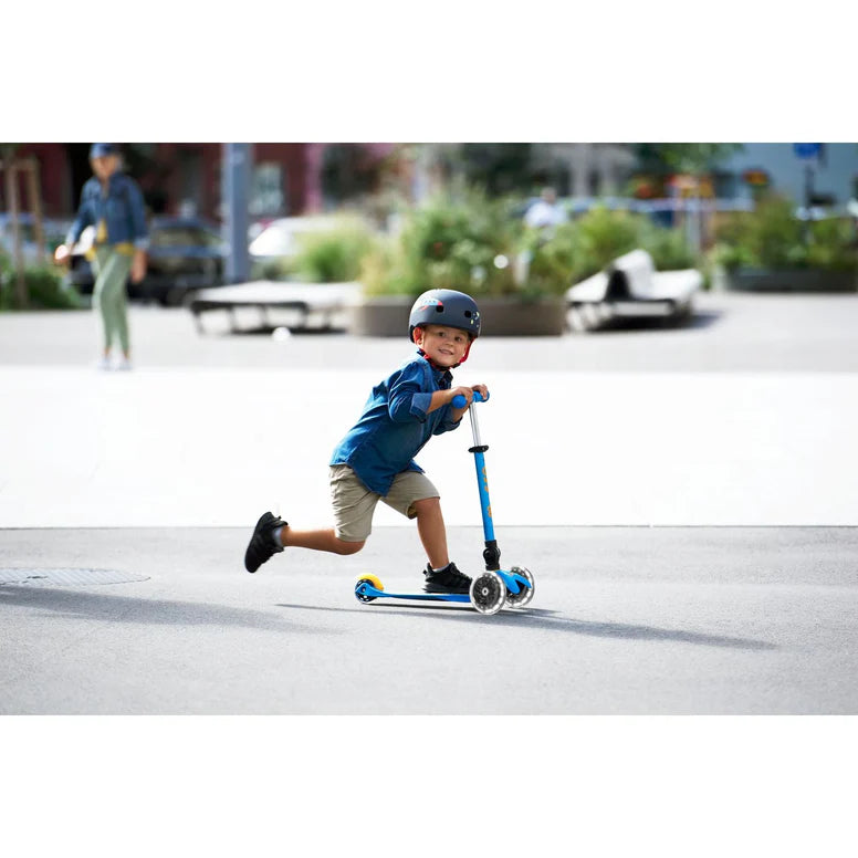 Micro Mini Deluxe Foldable LED Scooter, blue, as a child in a helmet rides excitedly down a sunny street.