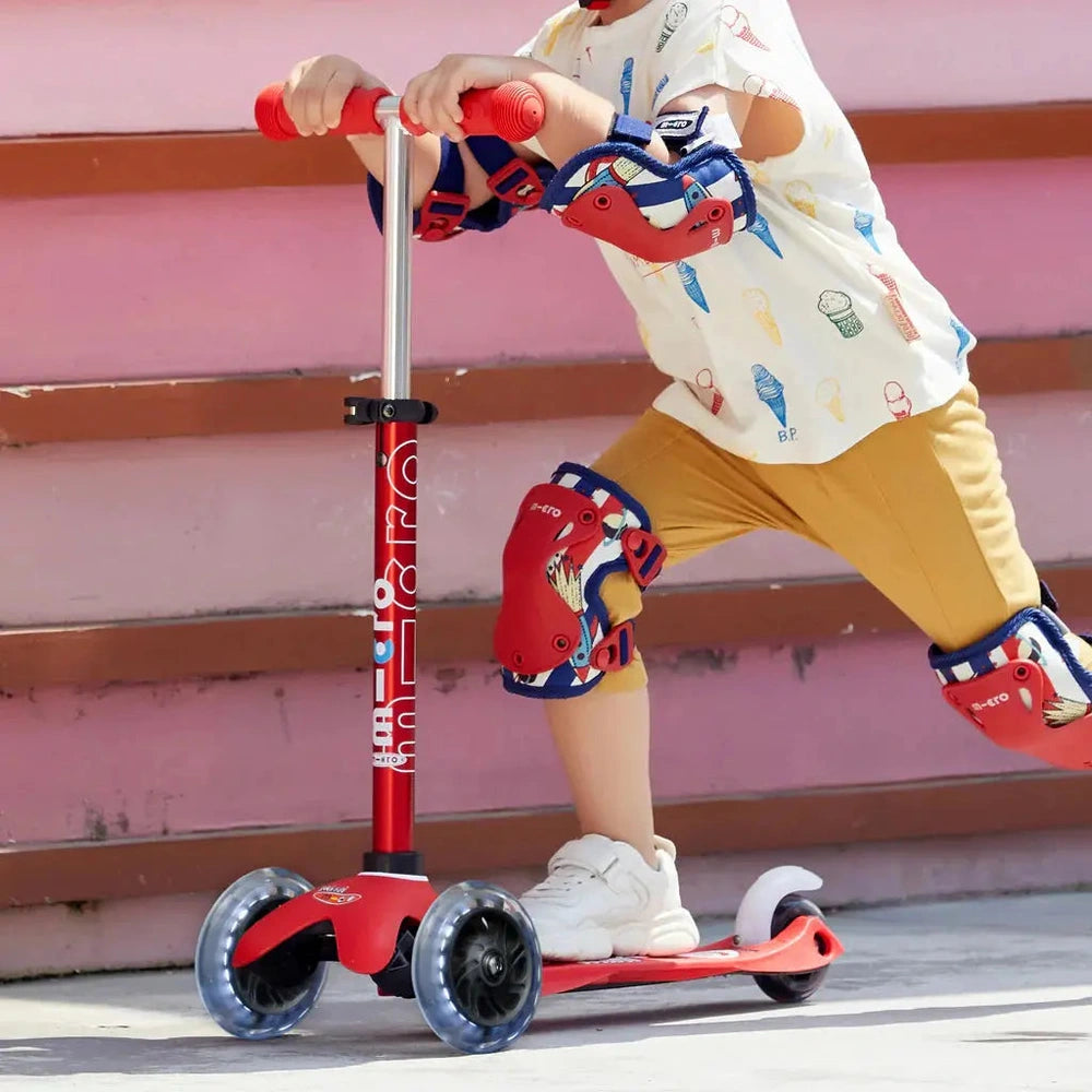 Micro Mini Deluxe LED Scooter, red with blue accents, child rides forward on scooter wearing knee/elbow pads.