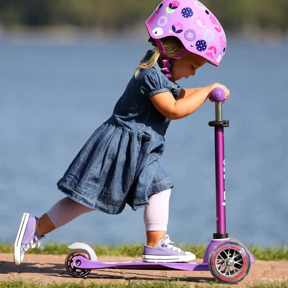 Micro Mini Deluxe Scooter for kids in purple with pink accents; child in a denim dress rides by a lake wearing a pink helmet.