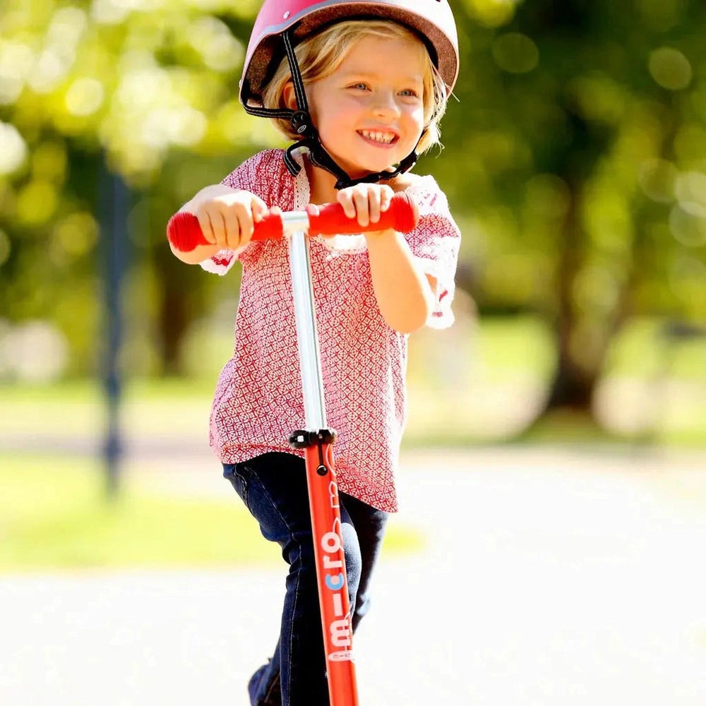 Micro Mini Deluxe Scooter, red frame, girl in pink helmet rides outdoors smiling.