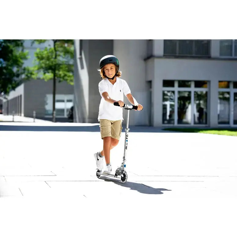 Micro Sprite LED Scooter, silver frame, ridden by a child outdoors with helmet in sunny urban plaza