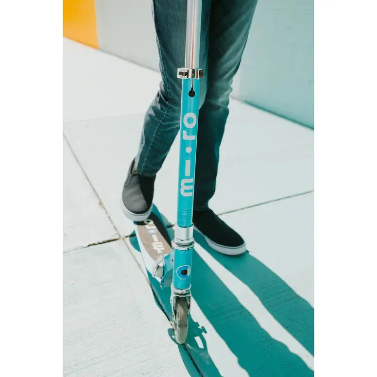 Micro Sprite LED Scooter in turquoise with chrome fittings; rider stands on deck, pushing along.