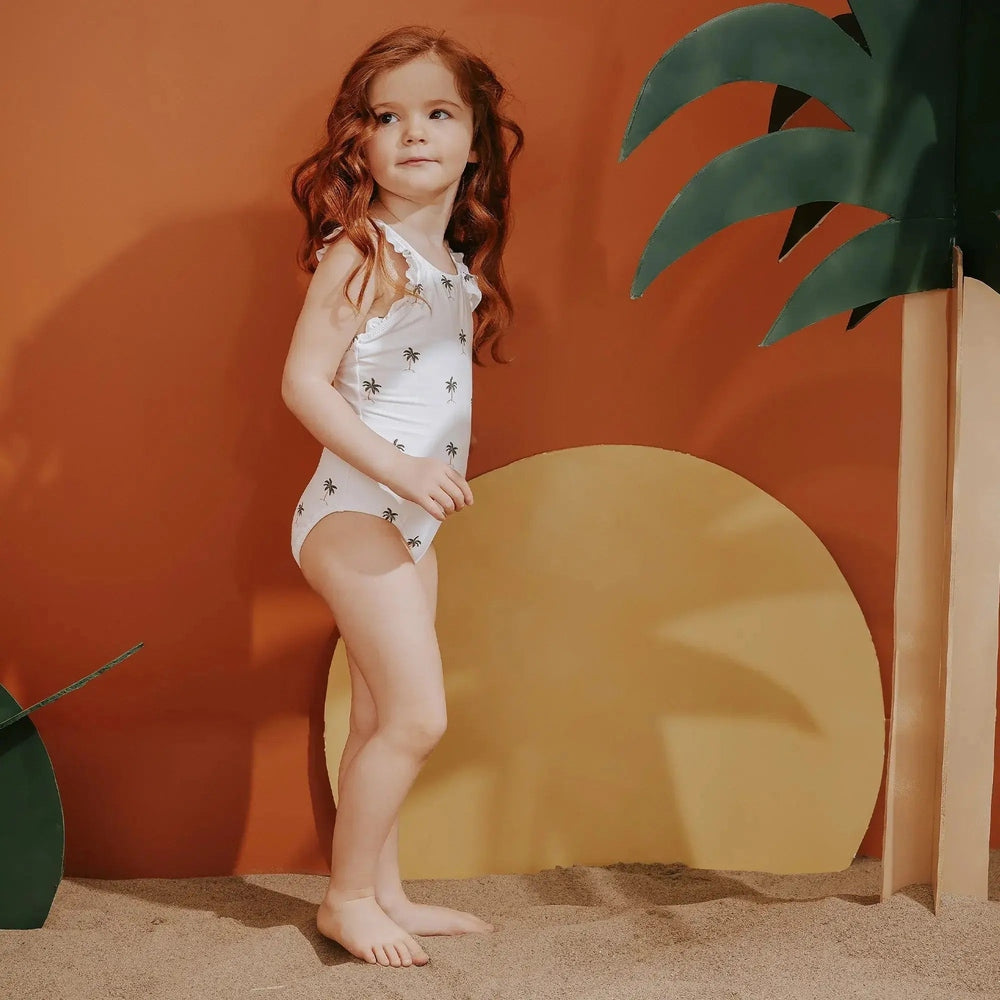 Miles The Label Ruffled Swimsuit Palm Trees for toddlers, white with palm print and frills, shown on a girl posing on sand.