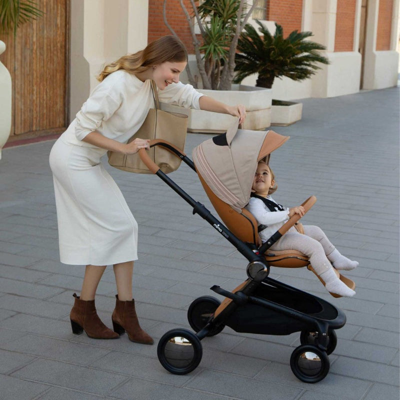 Mima Creo Stroller in beige with tan canopy and black frame as a parent pushes a child seated in the stroller.