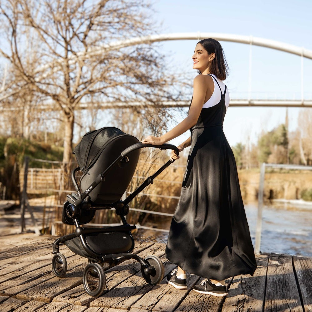 Mima Creo Stroller, black bassinet, pushed by a woman along a wooden boardwalk beside a river.