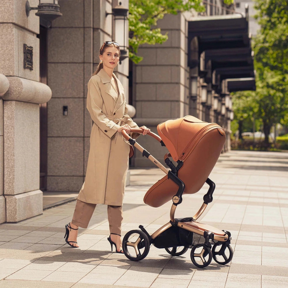 Mima Xari Max Stroller in tan, being pushed by a stylish woman along a city sidewalk.
