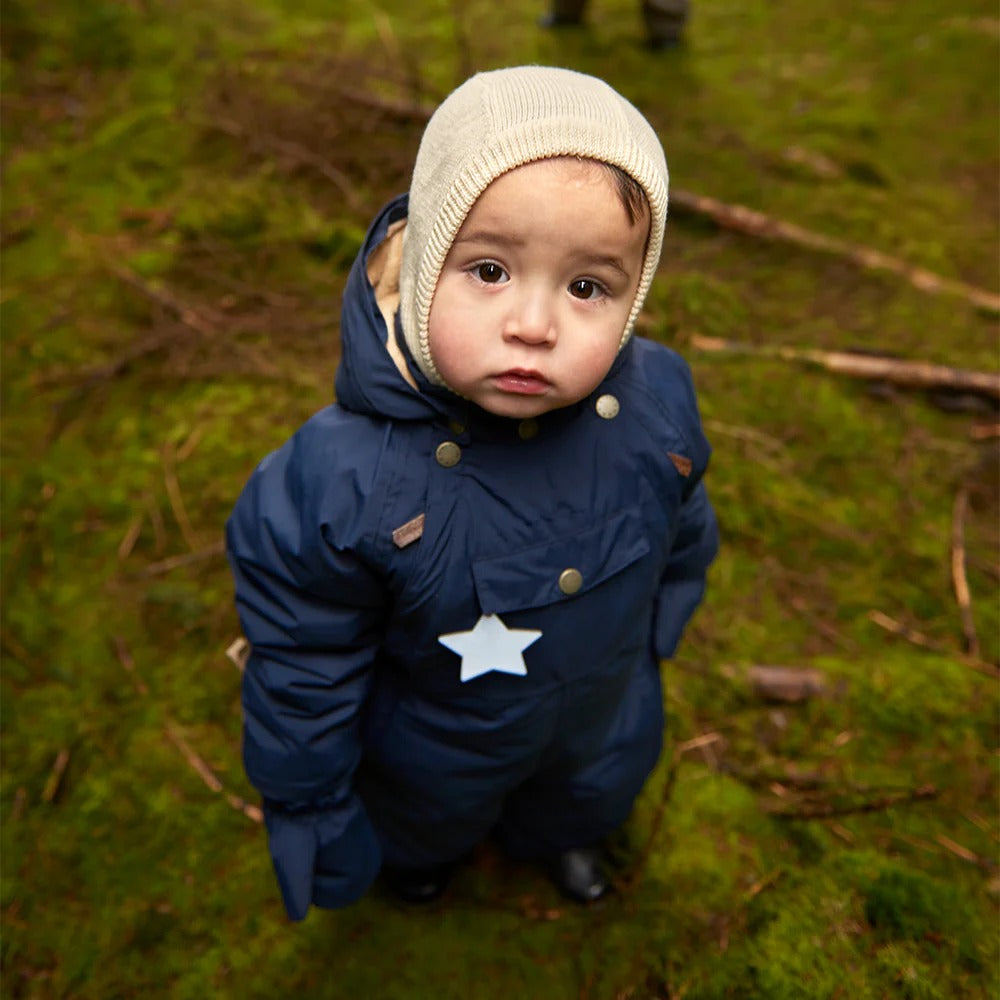 Baby in navy blue puffer snowsuit with cream angora knit hood and white star patch.