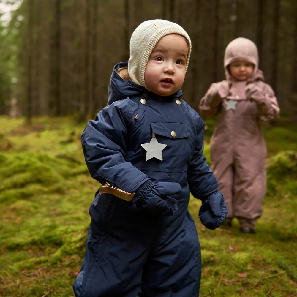 Baby in navy blue puffer snowsuit with cream angora-knit hood and star charm, forest setting.