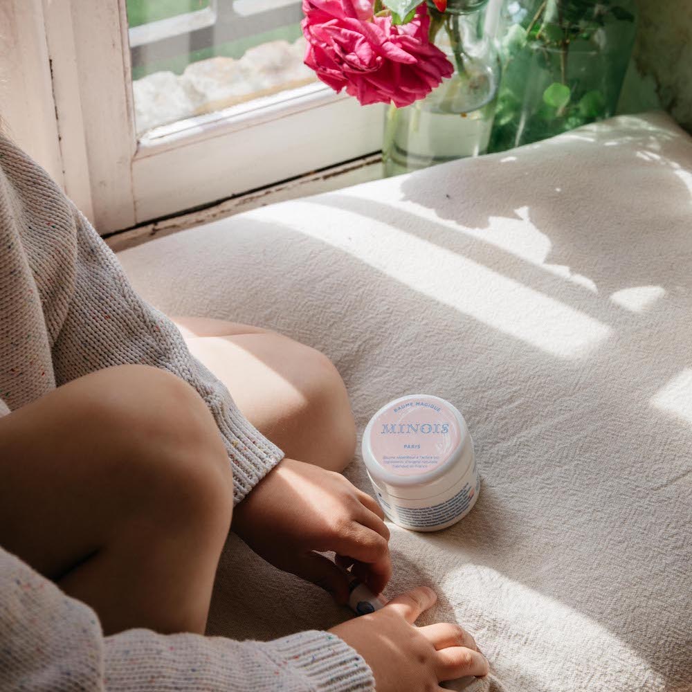 Minois Paris Magic Balm 50g jar on a beige couch with a child applying balm by a sunlit window.