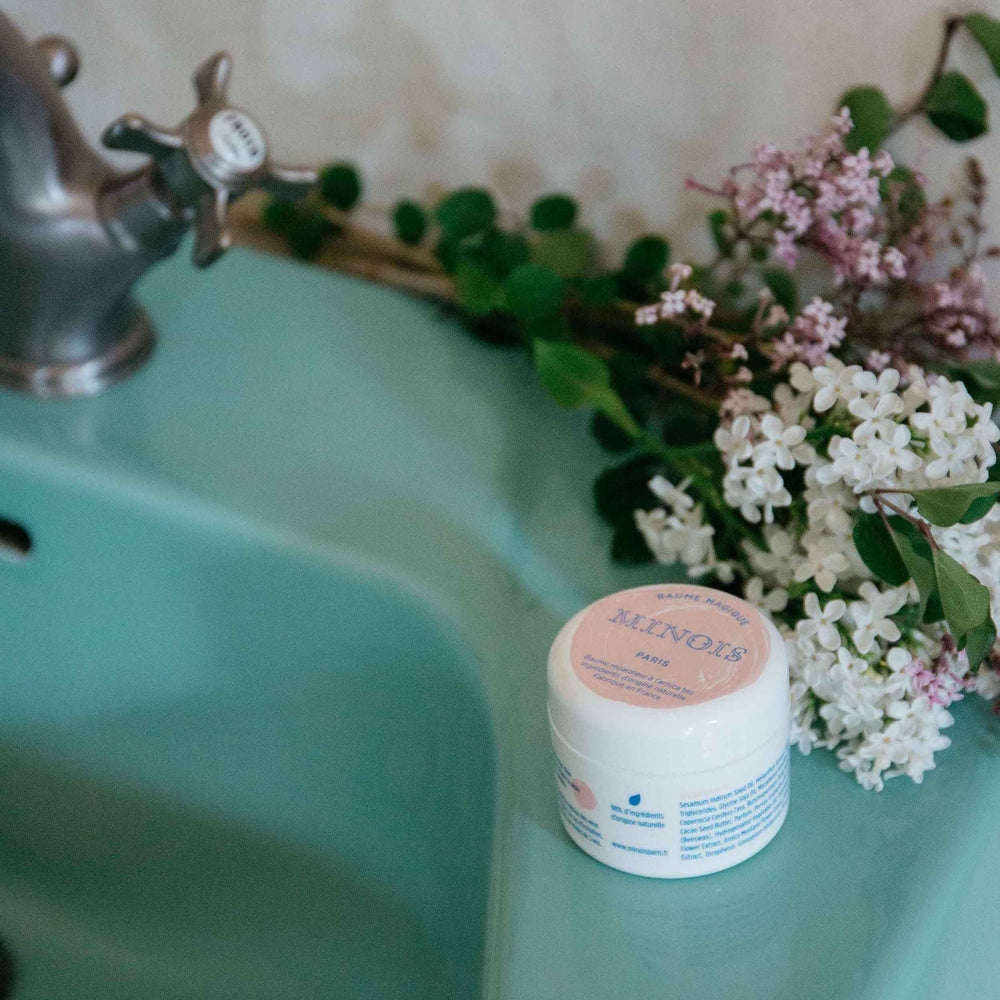 Minois Paris Magic Balm 50g jar on teal bathroom sink beside pink and white lilac bouquet.