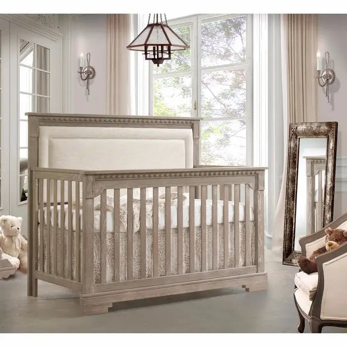 Natart Ithaca 5-In-1 Convertible Crib With Upholstered Headboard Panel in gray wood, shown in a bright nursery with slatted sides.