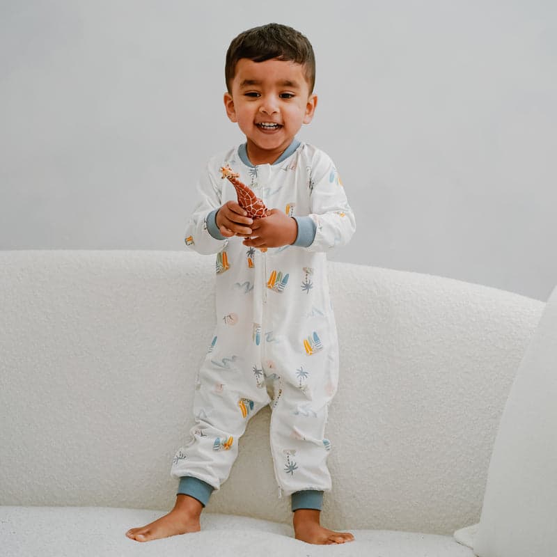 Nest Designs Bamboo Jersey One-Piece Zip Footless Sleeper Pastel Waves: toddler stands on a white sofa, holding a giraffe toy.
