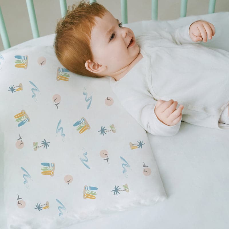 Nest Designs bamboo jersey toddler pillow with pastel waves pillowcase, supporting a sleeping baby on a white bed.