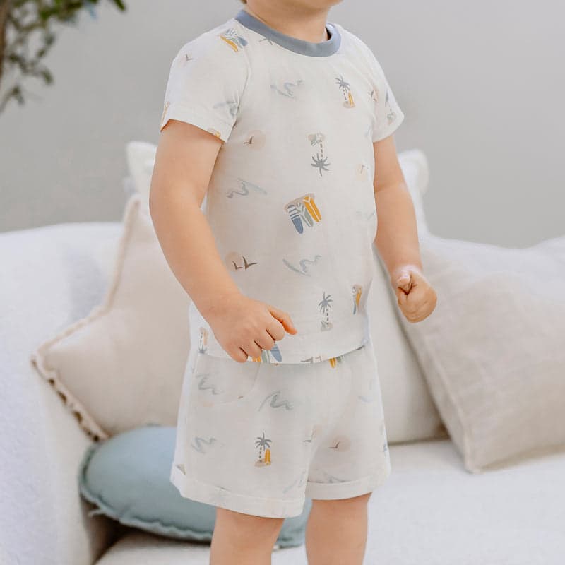 Nest Designs Bamboo Jersey Two-Piece Short Sleeve PJ Set Pastel Waves with palm tree print, worn by a toddler standing at home.
