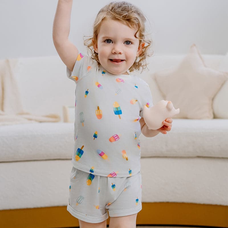 Nest Designs Bamboo Jersey Two-Piece Short Sleeve PJ Set Summer Pops, toddler in light gray with colorful popsicle print, raising arm.