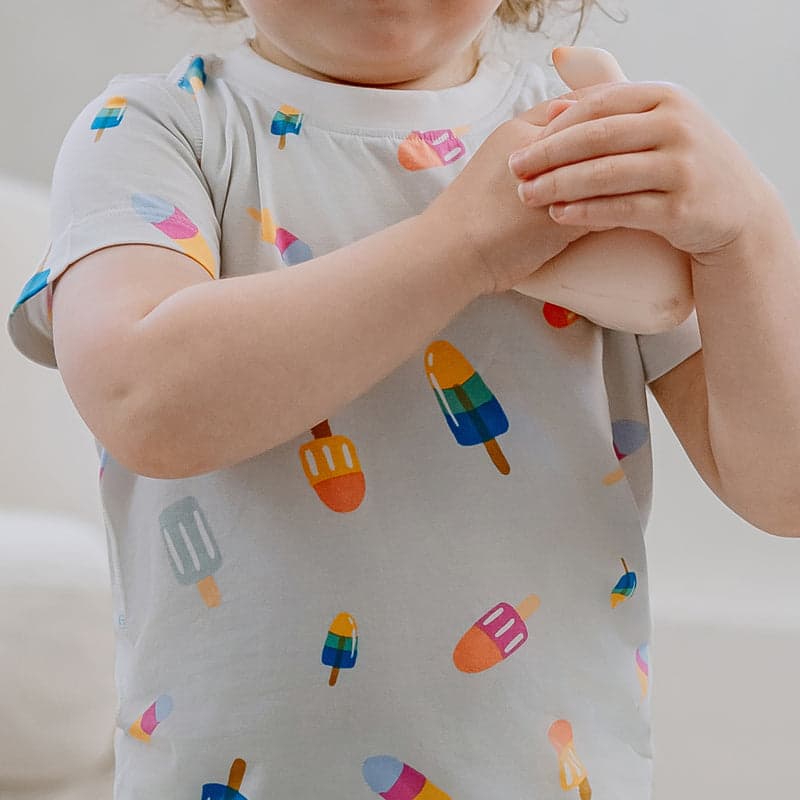 Nest Designs Bamboo Jersey Two-Piece Short Sleeve PJ Set Summer Pops; toddler in soft gray PJs with colorful popsicle print, hands clasped.