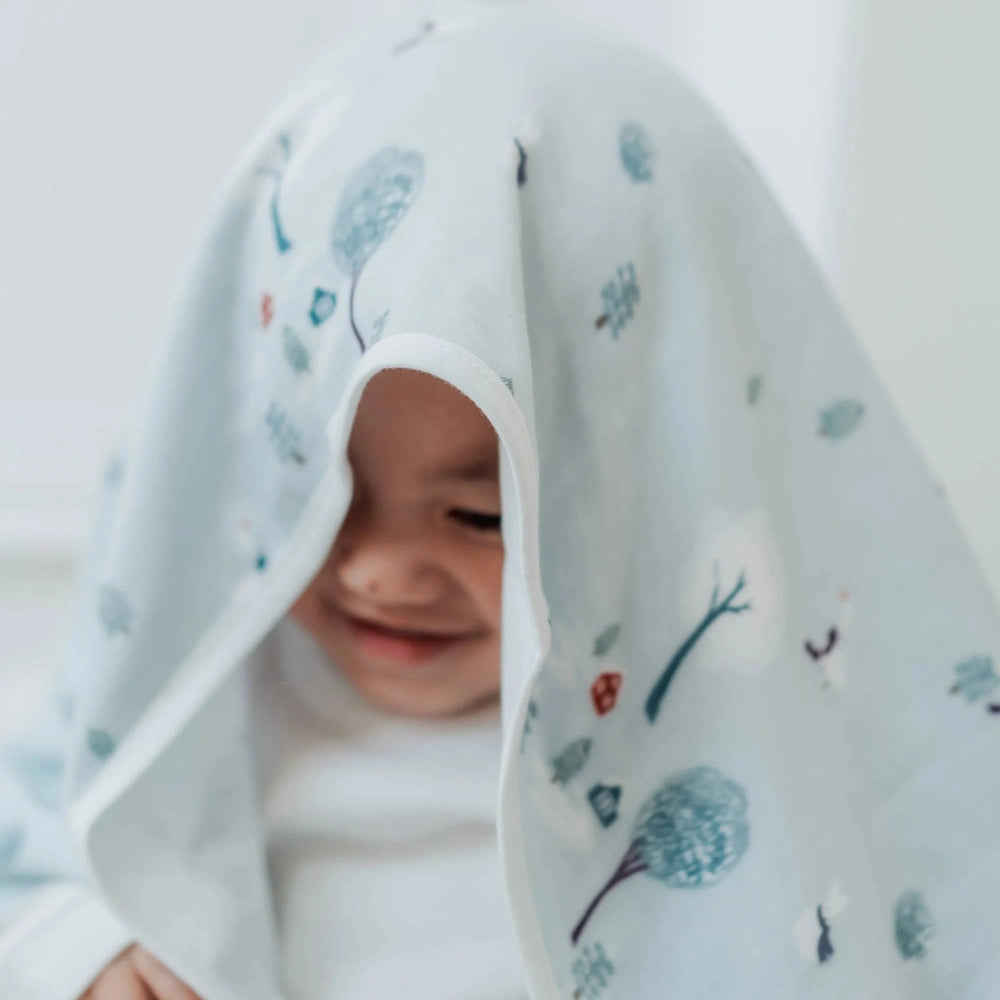 Nest Designs Bamboo Muslin Small Nesting Blanket 120cm x 120cm swaddles a smiling baby under a soft blue patterned cloth.