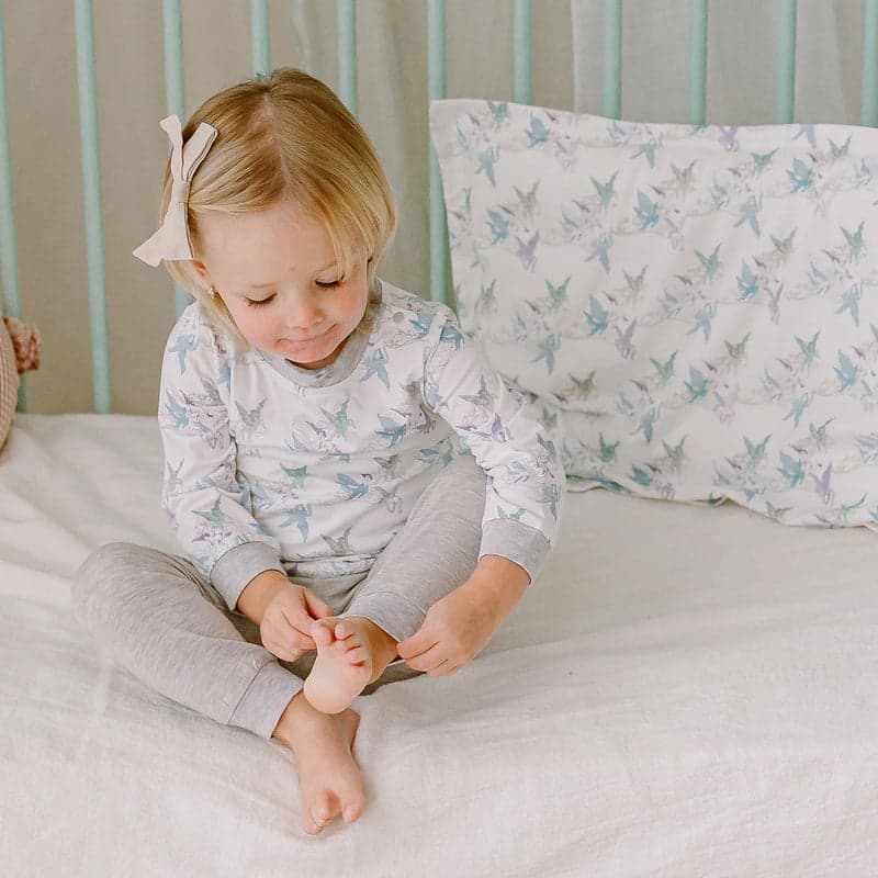 Nest Designs Bamboo Silk Toddler Pillow And Pillowcase Cloud Ponies; a toddler sits on a softly made bed with a cloud-ponies printed pillowcase.