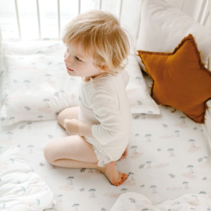 Nest Designs Bamboo Toddler Pillowcase Baby Baobabs, white with tiny trees, toddler sits upright on a bed.