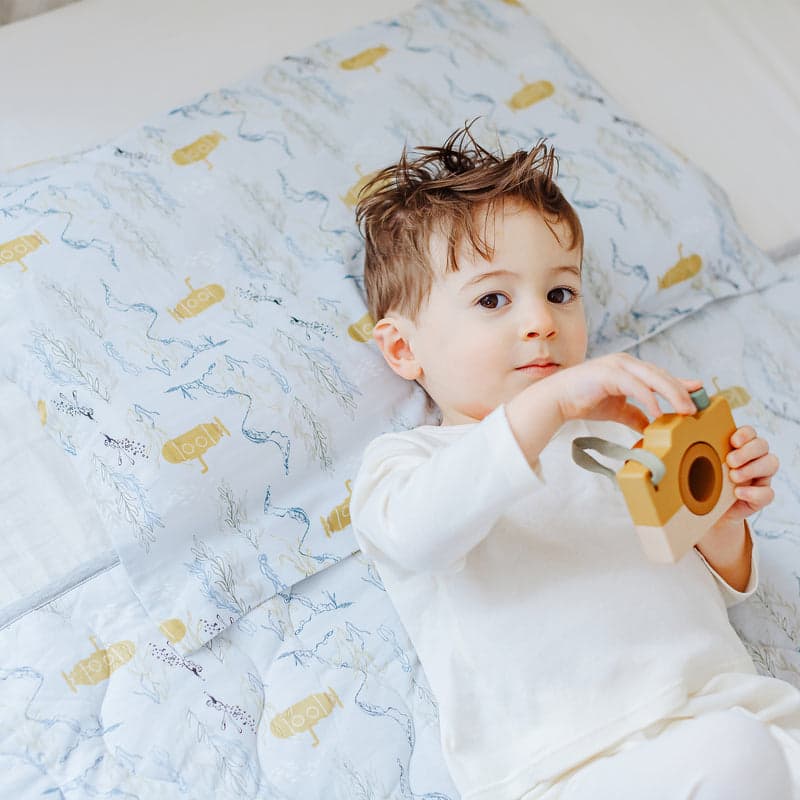 Nest Designs Bamboo Toddler Pillowcase Blub Blub Yellow Sub on light blue patterned bedding, child playing with a wooden toy camera