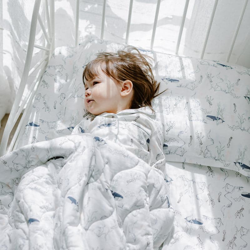 Nest Designs Bamboo Toddler Pillowcase Whale on white bedding with blue whale print; sleeping child tucked in.