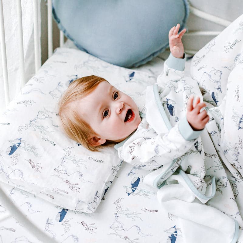 Nest Designs Bamboo Toddler Pillowcase Whale on white bedding; baby giggles with arms raised as the blue whale print shows.
