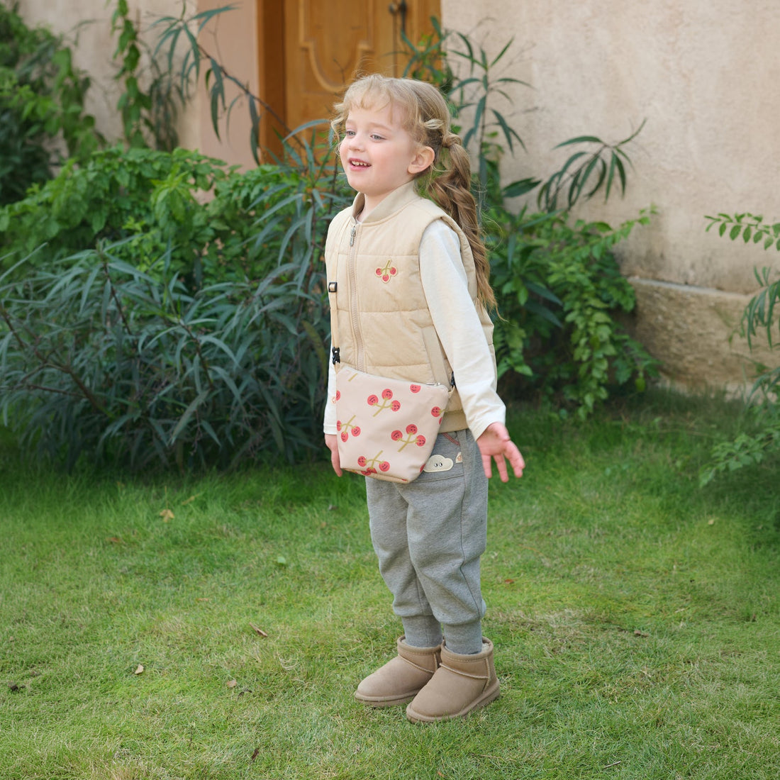 Child wears the Nest Designs Toast Biscuits & Vine Puffer Vest with grey pants and a cherry-print crossbody bag on grass