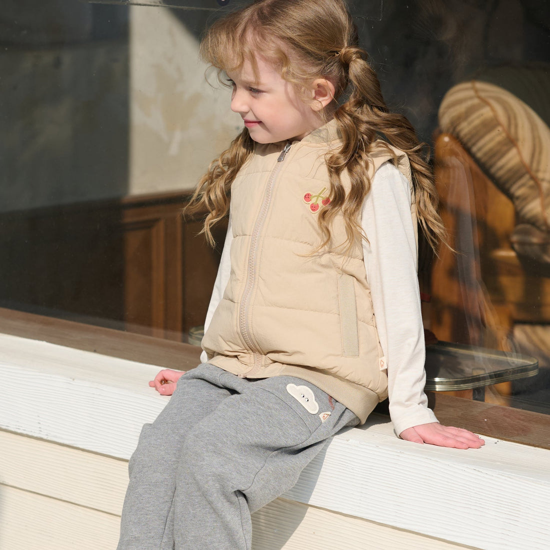 Child sits on a shop ledge in the Nest Designs Toast Biscuits & Vine Puffer Vest layered over a cream top and grey pants