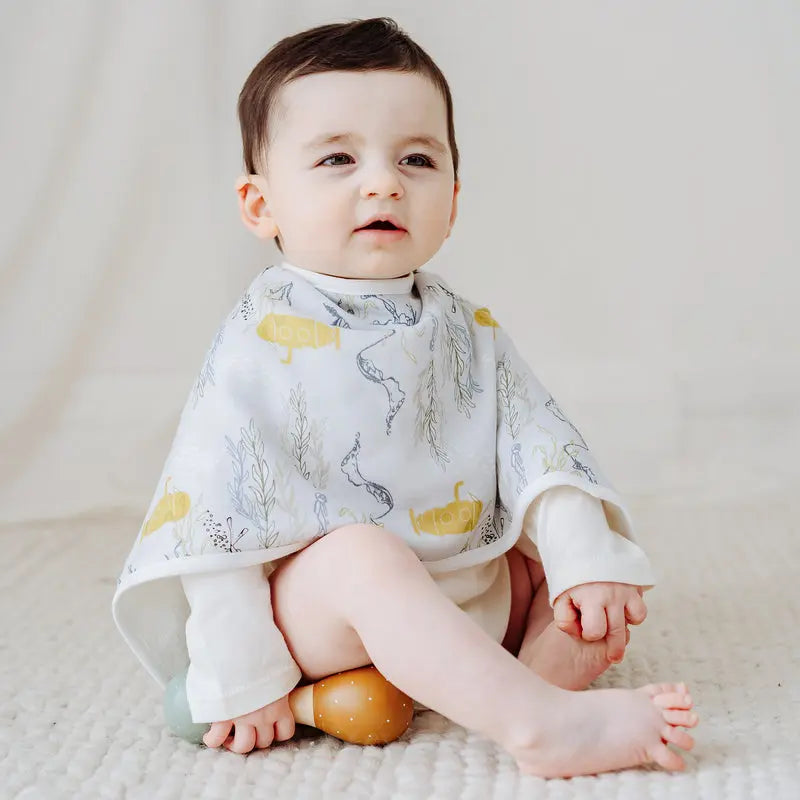 Nest Designs Burp'N Bib (2 Pack) ivory bibs with yellow-gray animal print, shown on a baby sitting on a cream rug
