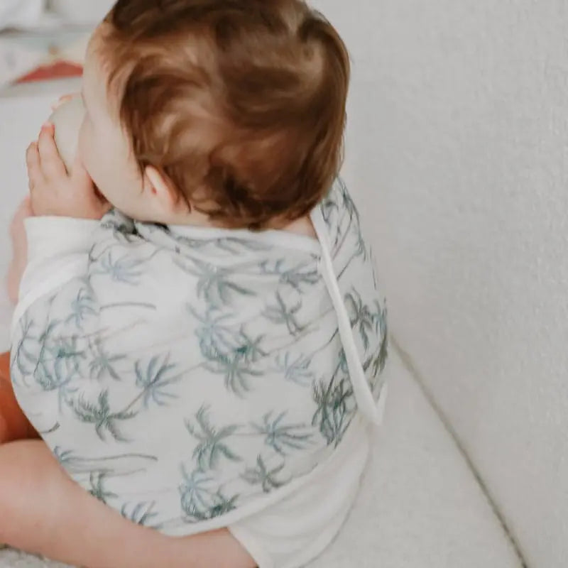 Nest Designs Burp'N Bib (2 Pack) white palm-tree print; baby drinks from a bottle with the neck strap visible.