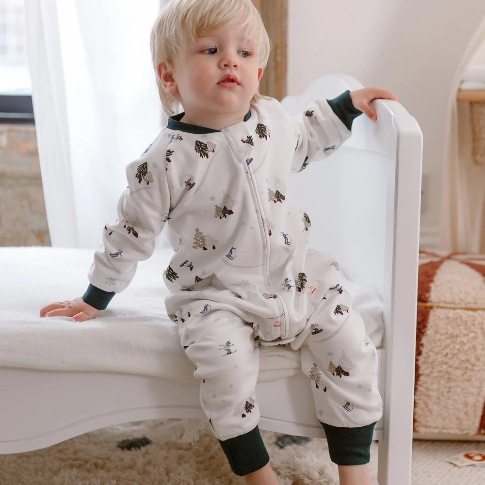 Nest Designs Cotton Long Sleeve Footed Sleep Bag 1.0 Tog; white with forest print, dark green cuffs, toddler sits on bed.
