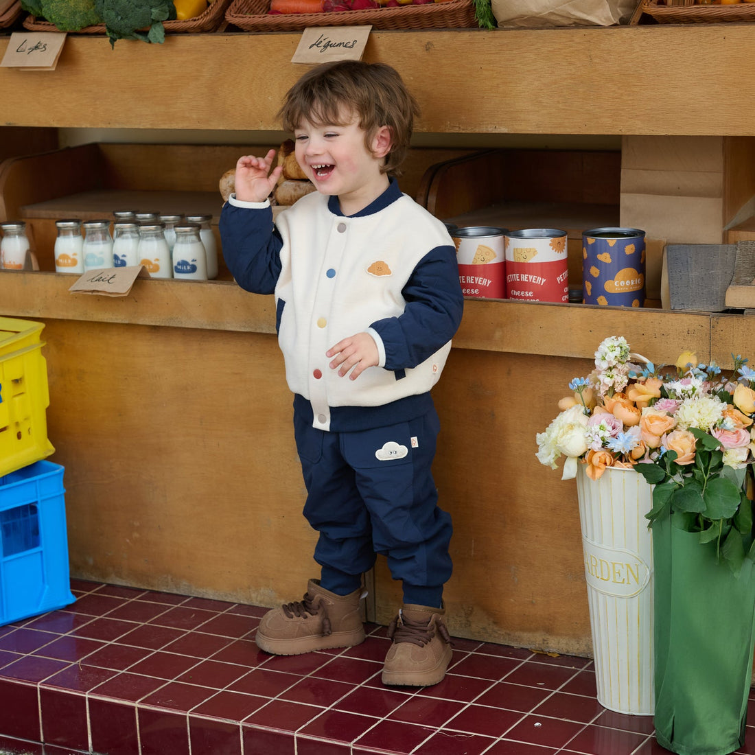 Child wears the Nest Designs Blueberry Cozy Croissant Fleece Jacket with navy pants while smiling at a produce stand