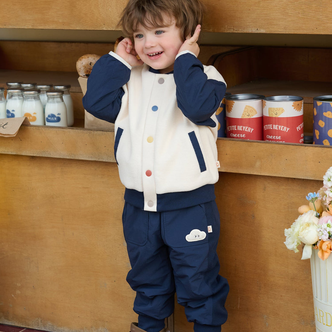 Child in the Nest Designs Blueberry Cozy Croissant Fleece Jacket and navy pants poses with hands by the ears at a market