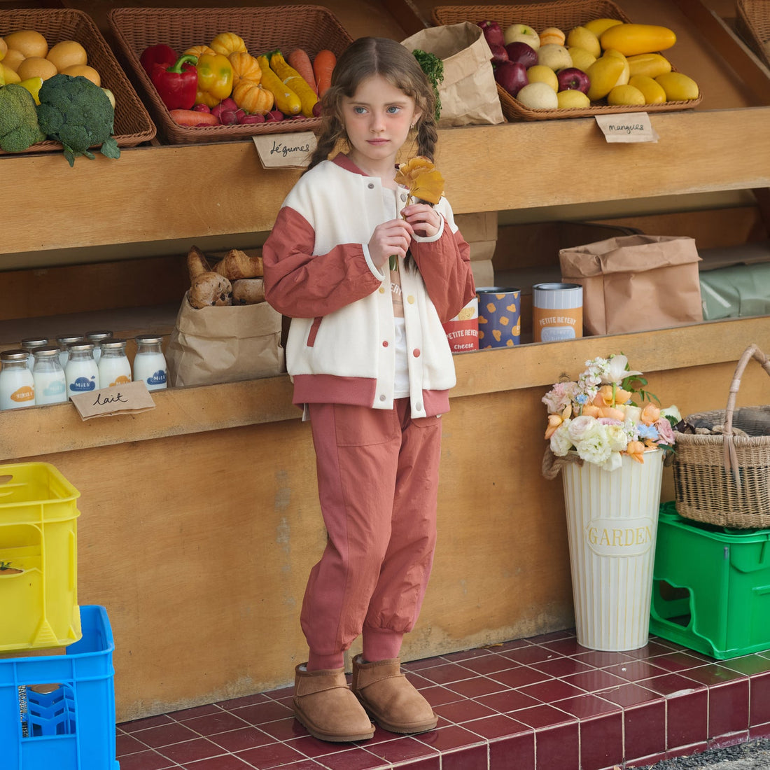 Child wears the Nest Designs Redwood Burl Cozy Croissant Fleece Jacket with matching pants while holding a flower at a produce stall