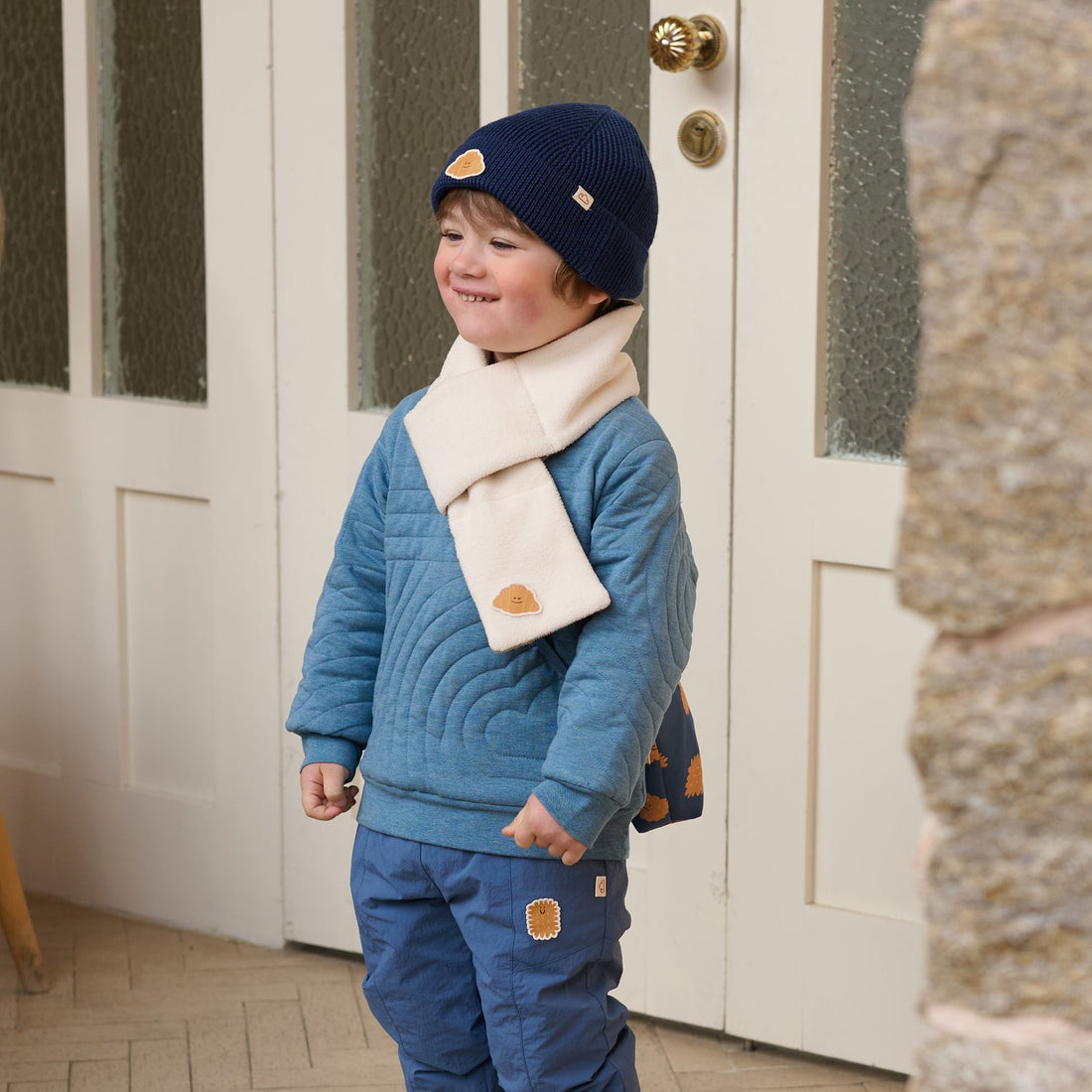 Nest Designs Cozy Croissant reversible scarf in Cloud cream worn outside a doorway with a blue quilted outfit