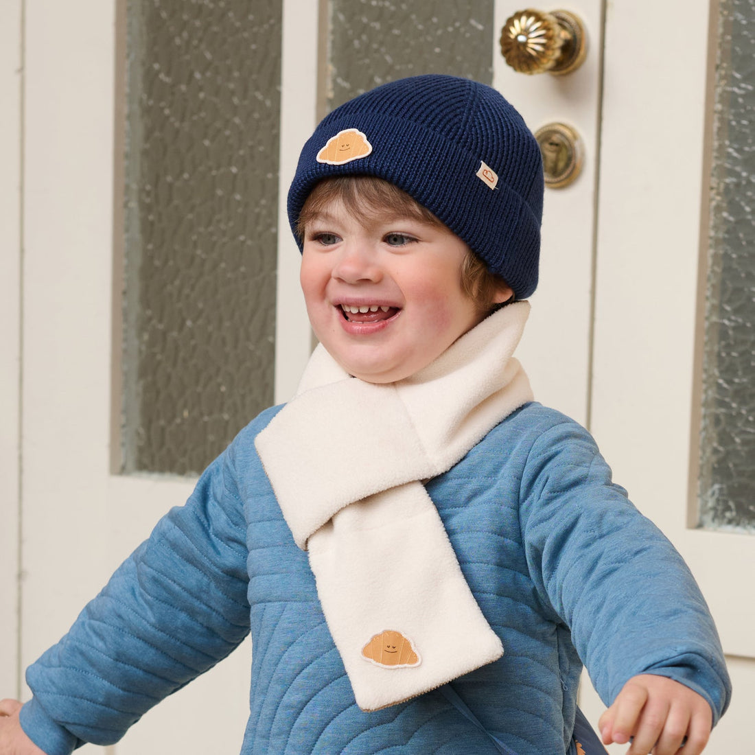 Nest Designs Cozy Croissant reversible scarf in Cloud cream worn by a smiling child moving with arms raised
