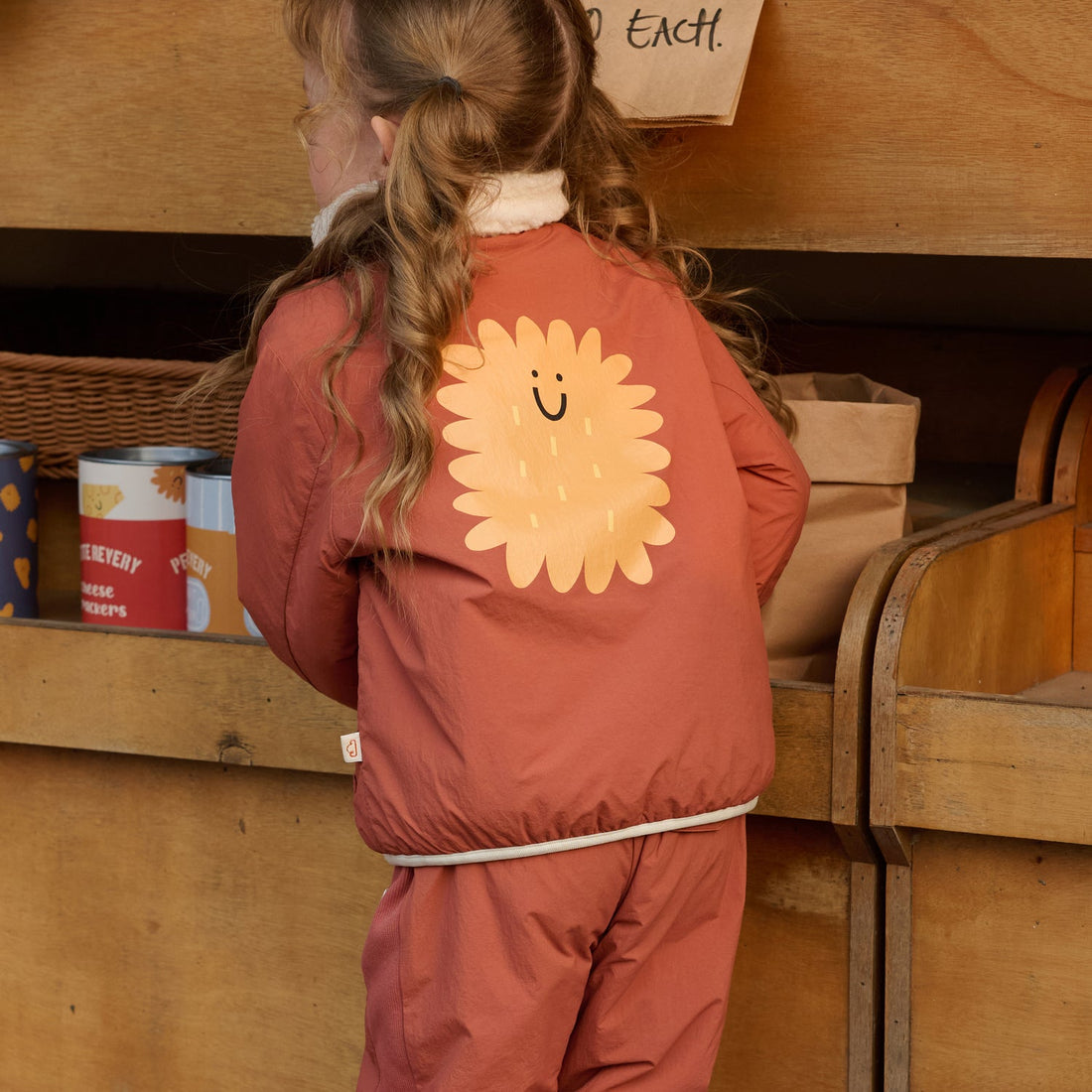 Nest Designs Cozy Crumbs reversible jacket in Redwood Burl rust, rear worn view with large smiling biscuit graphic at a market