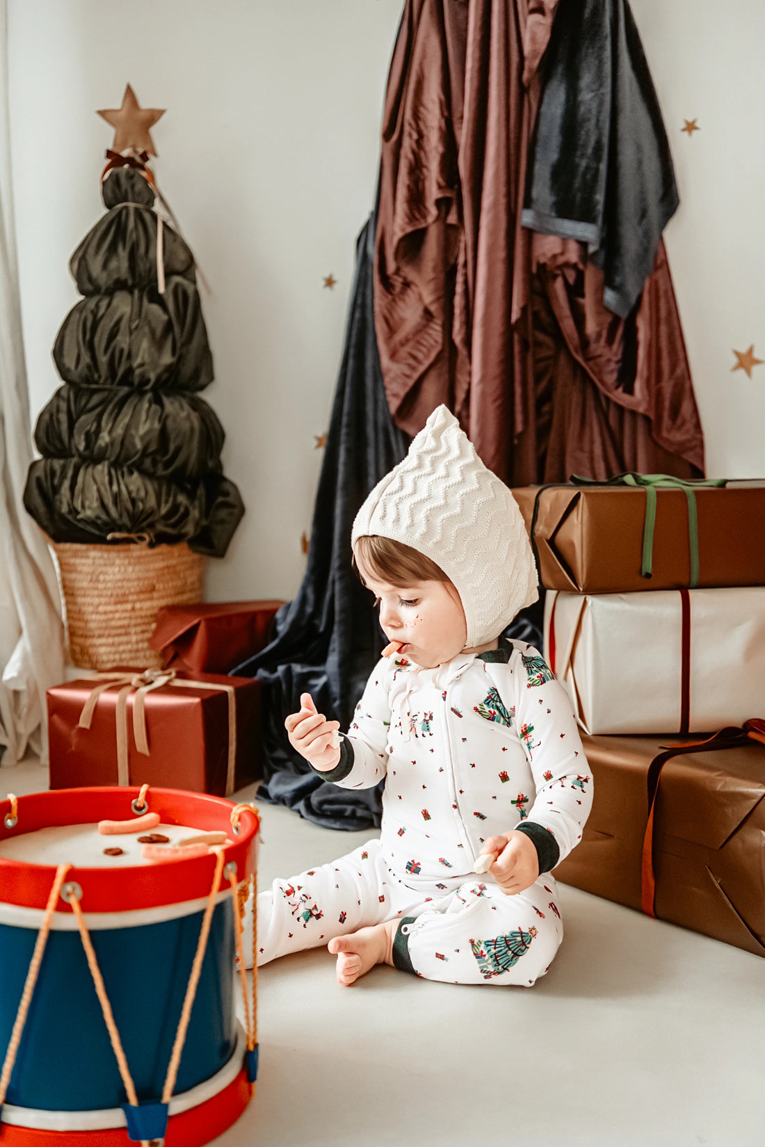 Nest Designs Santas Elves: child seated beside a toy drum in the footless sleeper; styling accessories are not included