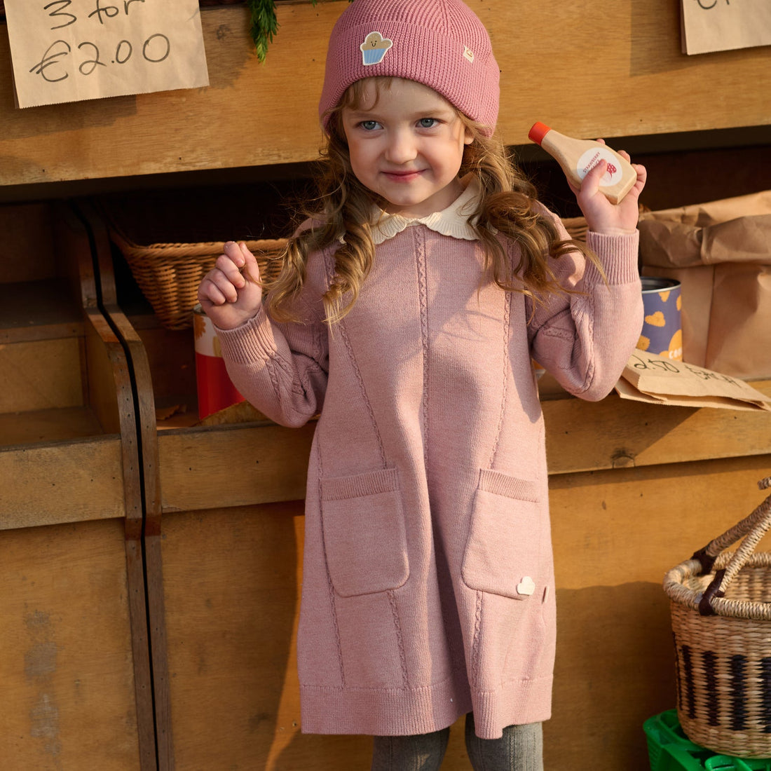 Nest Designs Rose Cable Knit Dress in Old Rose worn front-facing by a child holding a small item at a market