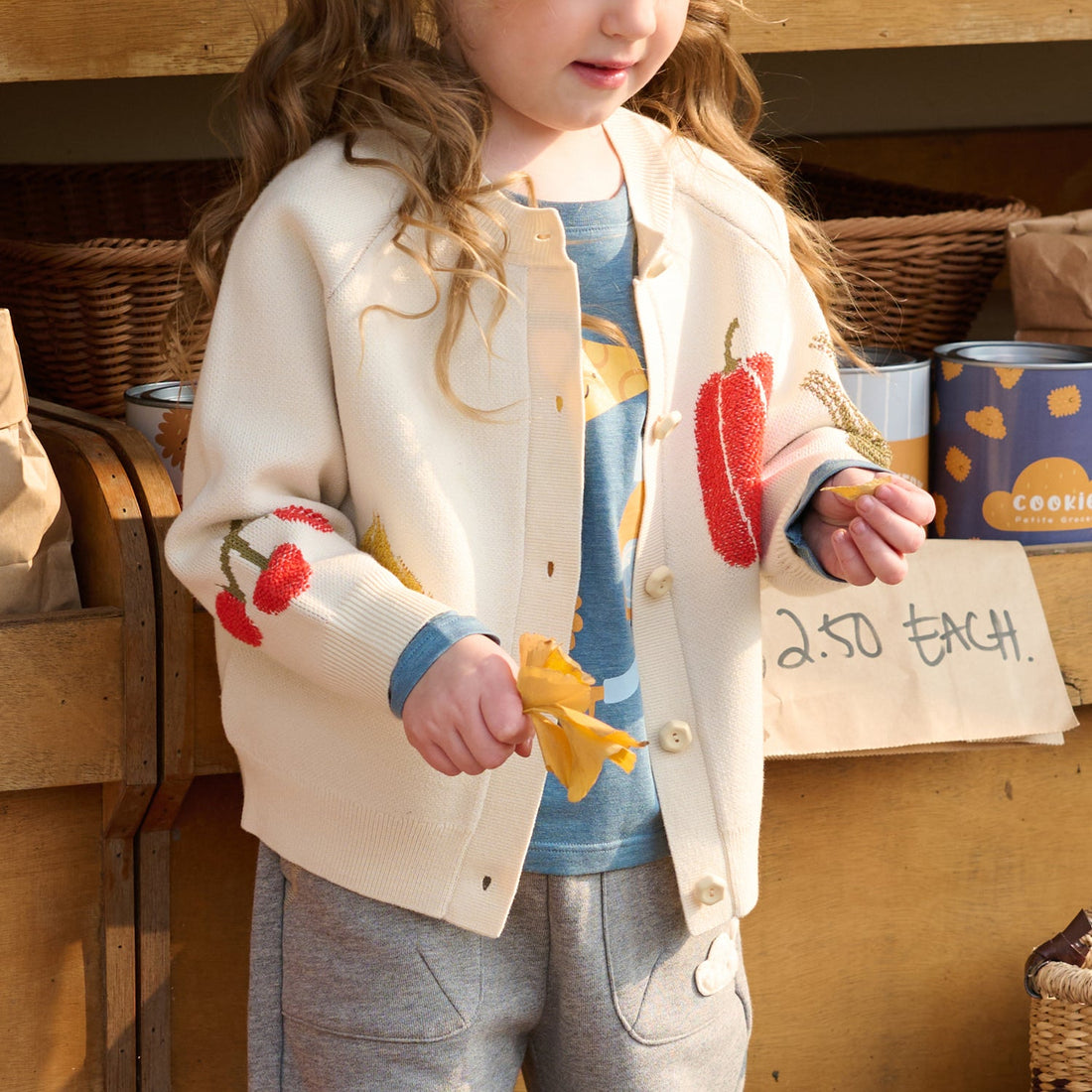 Nest Designs Jet Stream cream knit cardigan worn open over blue while a child holds a yellow pepper