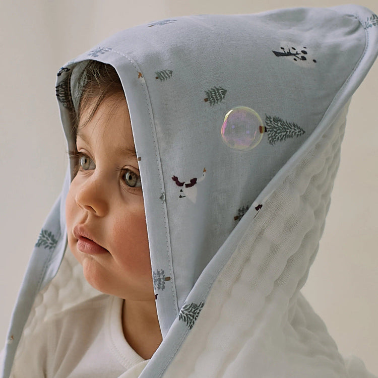 Nest Designs Organic Cotton 9-Layer Hooded Baby Bath Towel Snowy Snuggles; baby wears a light-blue hooded towel with trees.